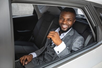rich african american young businessman smiling through open car window. cheerful handsome black man in stylish suit going to business trip by car, sitting on back seat and looking through window
