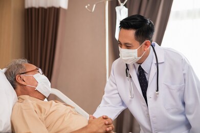 asian male doctor consoling and talking with elderly man patient during medical visit at hospital ward. medical professional  reassuring senior patient while recovery at hospital.