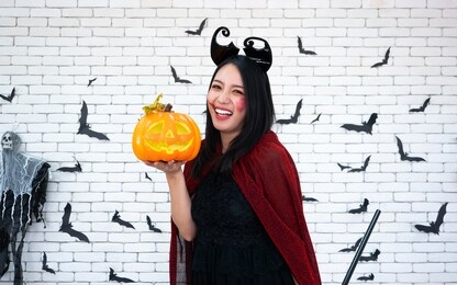 asian pretty woman smile and hold plastic pumpkin craft while wearing witch fashion costume portrait with decoration black bat on white brick wall background for happy halloween festival party theme