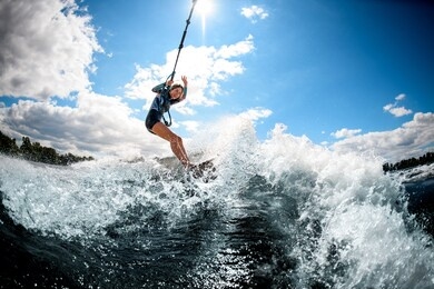 young active cheerful woman riding on the wake surf holding rope of motorboat and water splashing around.