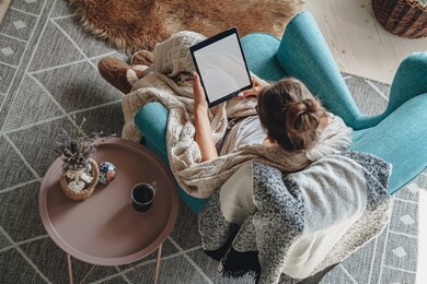 young woman sitting in cozy armchair, with warm blanket, using tablet