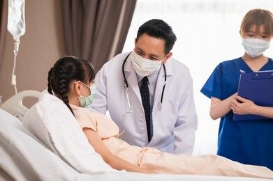asian male doctor consoling and talking with recovering girl patient during medical visit at hospital ward  with female nurse standing in background. medical workers visiting a girl patient.