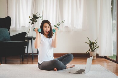 happy and excited asian woman holding dumbbells in both hand feeling ready to exercises workout at home with laptop 