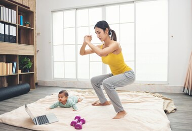 sporty asian woman in yellow sportswear is exercise on the floor with daughter baby, dumbbells and using a laptop. young mother and infant make squat pose at home in the living room.