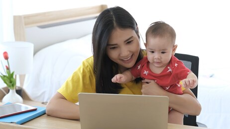 beautiful business asian mom is using a laptop and smiling while spending time with her cute baby at home. young mother on maternity leave trying to freelance by the desk with toddler child. 