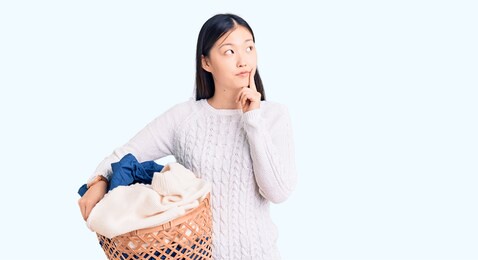 young beautiful chinese woman holding laundry basket with clothes serious face thinking about question with hand on chin, thoughtful about confusing idea 