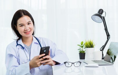 asian doctor young beautiful woman smiling using working or holding with smart mobile phone and laptop computer at hospital desk office, technology healthcare medical concept