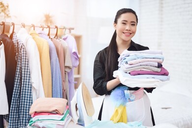 young asian maid puts ironed clothes in dressing room. asian woman smiling and holding ironed things in her hands.