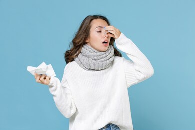 sick young woman in white sweater scarf hold napkin put hand on nose keeping eyes closed isolated on blue background studio portrait. healthy lifestyle ill sick disease treatment cold season concept