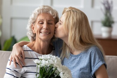i love you so much, my dear. affectionate millennial daughter greeting happy senior elderly mom with mothers day, happy grandma receiving tender kiss and flowers with closed eyes from grown grandkid