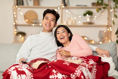 vietnamese couple watching tv on christmas evening enjoying movies and popcorn sitting on sofa at home. japanese family covered with blanket watches xmas television shows during new year holidays