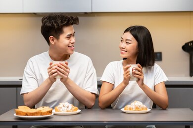 lovely asian couple having breakfast sitting in kitchen, enjoying coffee and croissants on weekend morning. chinese family bonding having romantic brunch, eating pastry and drinking tea at home.