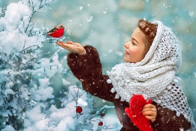 photo as a postcard in vintage style depicting a small girl in a white downy shawl and red mittens, smiling and feeding a bullfinch bird from her hand, while white fluffy snow is falling around.