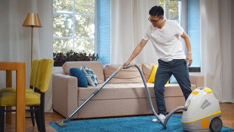 young asian man cleaning house with vacuum cleaner. portrait of handsome korean guy vacuuming blue carpet in modern living room doing household.
