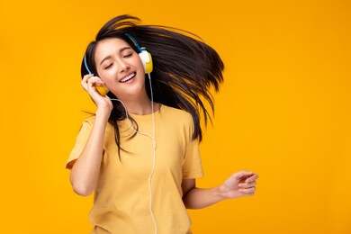happy young asian woman listening music in headphones and dancing on yellow background. charming girl get fun and enjoying life. attractive female enjoying her favorite song. copy space