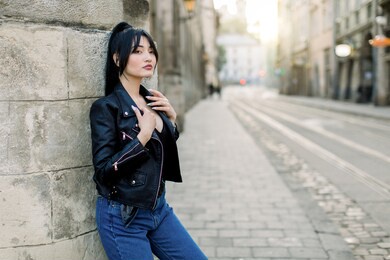 portrait of a beauty brunette asian fashion model in stylish leather jacket and denim jeans, posing near the wall of building. street of old city and pavement road on the background