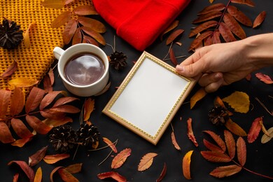 holding an empty photo frame in hand next to a cup of tea on a black background with autumn leaves, pine cones and needles, scarf and hat for cold weather