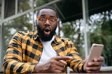 handsome african american man with emotional face shopping online, pointing finger on mobile phone screen. funny student studying online, watching training courses