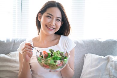 young happy asian woman eating healthy salad sitting on the sofa in living room.