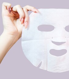female hands holds white cloth facial mask. over pink background