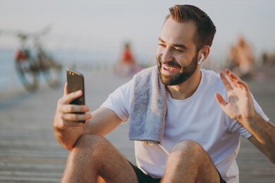 portrait funny young bearded fitness athletic strong man 20s in white t-shirt listening music with air pods doing selfie shot on mobile phone making video call resting at sunrise over the sea outdoors