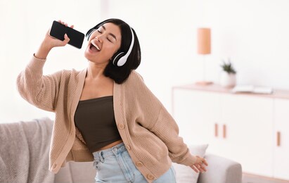 karaoke app. portrait of emotional asian woman singing her favorite song, dancing and using cellphone as mic, wearing wireless headphones. excited lady having fun at home in living room, copy space