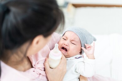 asian mother holding and feeding crying baby from milk bottle. portrait of cute newborn child being fed by mother on sofa in living room. mom giving nutrition food to kid drinking the milk at house.