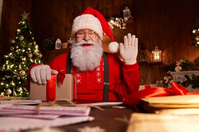 happy santa claus, saint nicholas holding letter looking to camera, talking to webcam video calling or recording happy new year, merry christmas greetings on xmas eve sitting at table late at home.