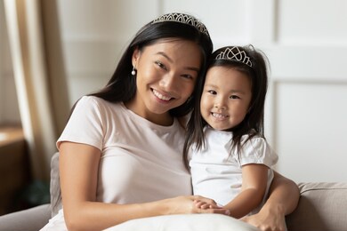 head shot portrait beautiful smiling asian mother and little daughter looking at camera, sitting on cozy couch at home, happy young mum hugging adorable child, posing for family photo together