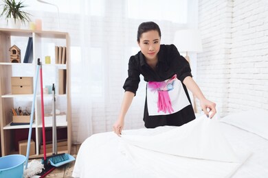 chinese housemaid woman is changing bed at house. house cleaning. asian woman in special uniform looks at the camera while making a bed.