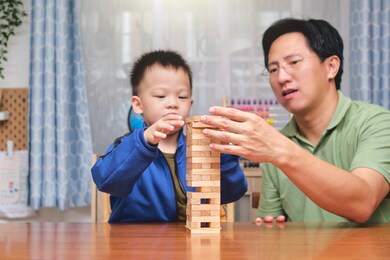 happy father and cute little asian toddler boy child excited with wooden block jenga game, dad and son spending quality time together playing tower games indoor at home - selective focus at dad's hand