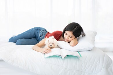 a young asian woman lounging with a maltese dog on a white bed.