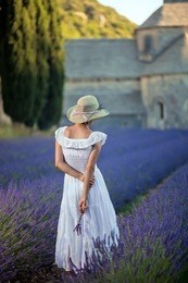 romantic lady in lavender field. an ancient monastery in background. wearing white dress and light green hat, holding some lavender twigs in her hand. relaxing and enjoying.