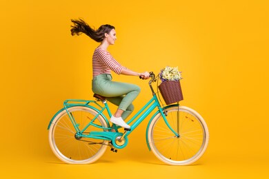 full length body size side profile photo of girl riding blue bicycle with basket of wild flowers isolated on vibrant yellow color background.