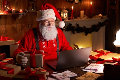 happy old santa claus wearing hat holding gift box using laptop computer sitting at workshop home table late on merry christmas eve. ecommerce website xmas time holiday online shopping e commerce sale