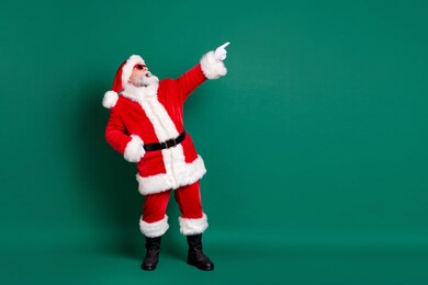 full length body size view of his he nice attractive handsome bearded cheerful cheery santa having fun celebratory festive day pointing copy space advert look isolated over green color background