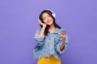 happy asian girl wearing headphones listening to music from mobile phone relaxing with eyes closed on isolated purple studio background