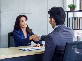 asian businesswoman and businessman wearing suit shaking hands in office. smiling female manager sitting at desk in office handshaking with young male employee after working together.