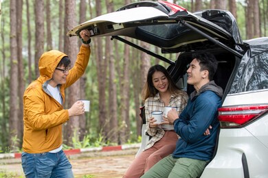 happy asian man and woman sitting in open car trunk at natural park. group of friends enjoy talking and drinking coffee together in travel road trip. friendship and outdoor lifestyle vacation concept.