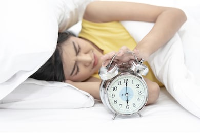 young beautiful asian woman holding alarm clock and trying to sleep under the pillow early in the morning