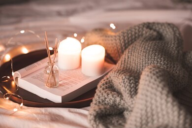 aroma sticks with liquid scented water in bottle with burning candles staying on open book and knitted textile in bed closeup over lights at background . winter holiday season. 