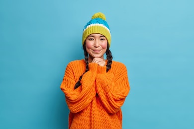 pretty asian girl keeps hands under chin gets red and shy as receives compliment from boyfriend wears warm knitted sweater and hat has charming gaze isolated over blue background. happy feelings