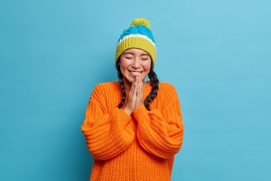 smiling teenage girl enjoys winter time keeps palms together and laughs happily with closed eyes being amused by friend dressed in knitwear isolated on blue background. positive emotions concept