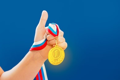 award and victory concept. woman hand raised with a thumb up sign, holding gold medal, blue background, copy space for text. first place in competition