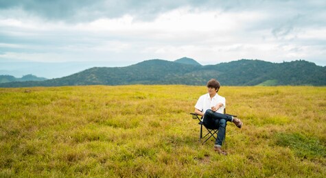 happy young asian man sitting alone on outdoor chair relaxing in beautiful nature of the mountain grass field in autumn day. loneliness handsome guy enjoy with outdoor lifestyle on holiday vacation