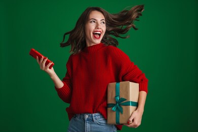 beautiful excited girl screaming while posing with christmas gift and cellphone isolated over green background