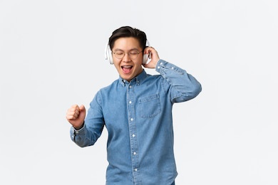 lifestyle, leisure and technology concept. happy dancing asian man in glasses, listening music wireless headphones, singing along, having fun with new album on streaming platform