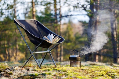 outdoor camping equipment close-up. stove with kettle, tourist folding chair with a book in the forest. the background is blurred.