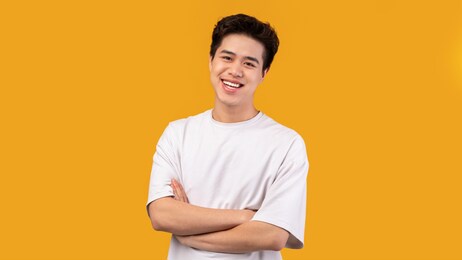 confident person. portrait of smiling asian guy with folded arms looking at camera, wearing white shirt, posing isolated over orange studio background. happy casual male teenage model laughing