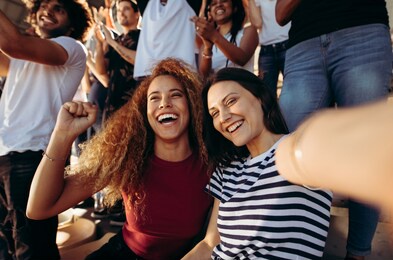 excited female spectators making a selfie while cheering their sports team from a stadium. cheerful men and woman fans taking selfie while watching match in stadium.